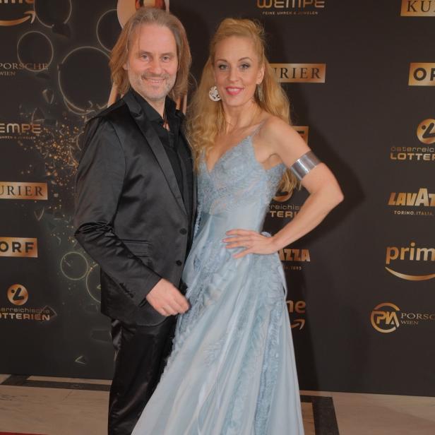 Ein Mann im schwarzen Anzug und eine Frau im hellblauen Abendkleid posieren gemeinsam auf einem roten Teppich.