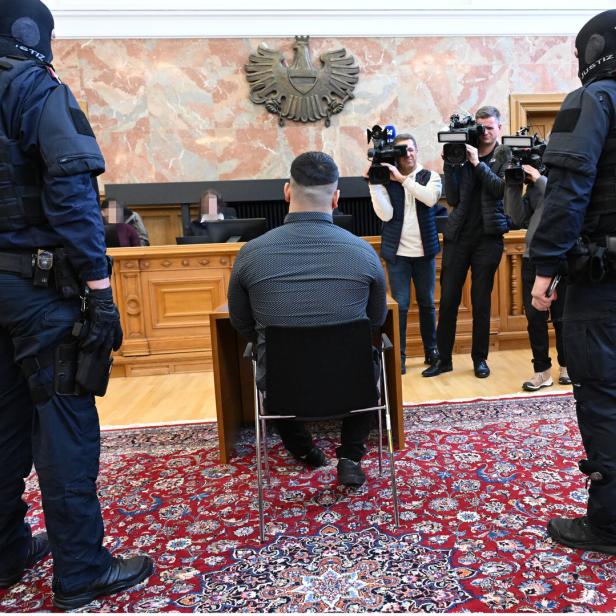 Mann sitzt mit dem Rücken zur Kamera in einem Gerichtssaal, flankiert von Justizwache