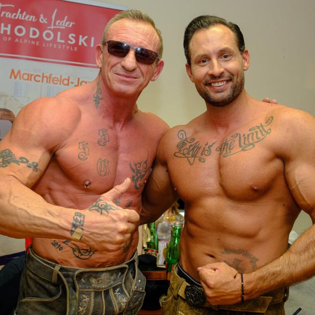 Zwei muskulöse, tätowierte Männer posieren oberkörperfrei und lächeln, einer zeigt den Daumen nach oben.