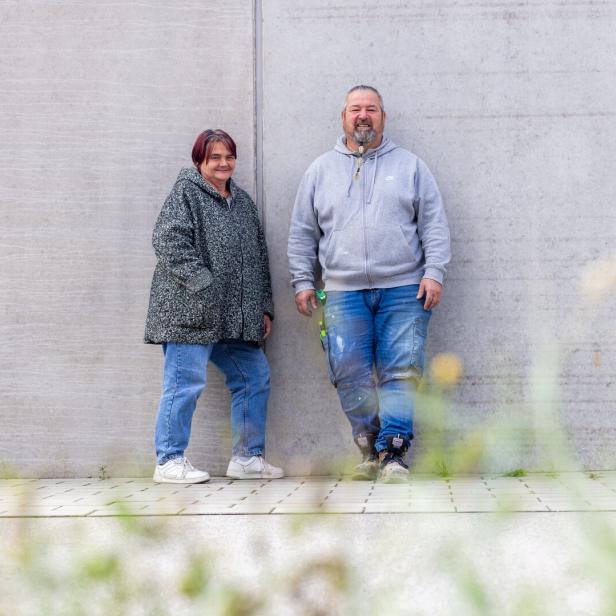 Zwei Personen stehen entspannt nebeneinander an einer grauen Betonwand, im Vordergrund unscharfe Gräser.