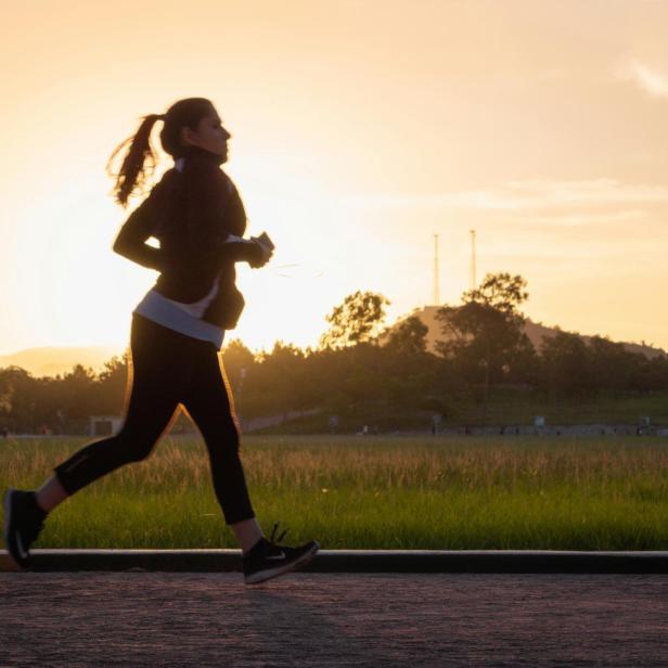 Silhouette einer joggenden Frau mit Pferdeschwanz, Sportkleidung und Laufschuhen vor Sonnenuntergang und Bäumen.
