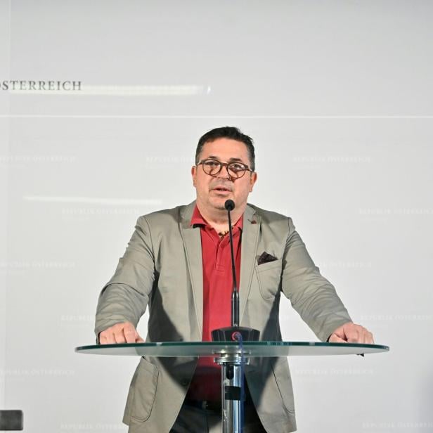 Ein Mann mit Brille spricht an einem Rednerpult vor einer Wand mit dem Logo und Schriftzug des österreichischen Parlaments.