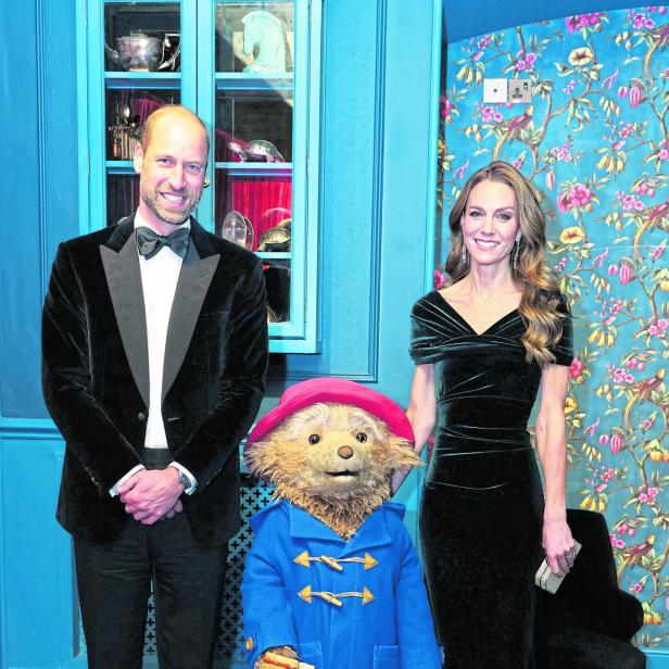 Prinz William, Kate und Paddington Bär posieren vor einer floralen Tapete.