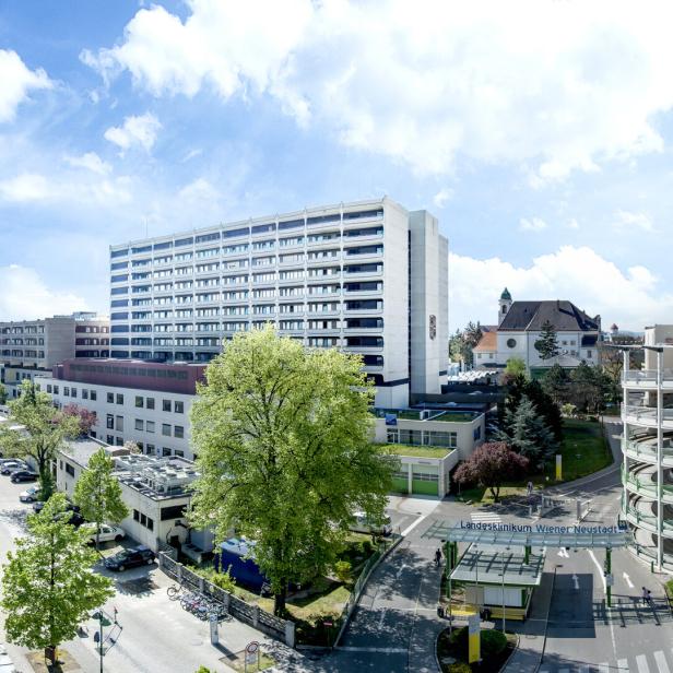 Mehrstöckiges Krankenhausgebäude mit Parkhaus, Bäumen und Zufahrt bei sonnigem Wetter.