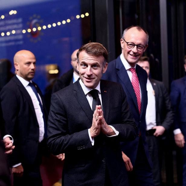 Emmanuel Macron mit gefalteten Händen, begleitet von Friedrich Merz und weiteren Personen.