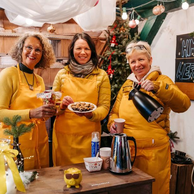 Drei Frauen in gelben Schürzen bieten Gebäck und Getränke an einem festlich geschmückten Stand an.