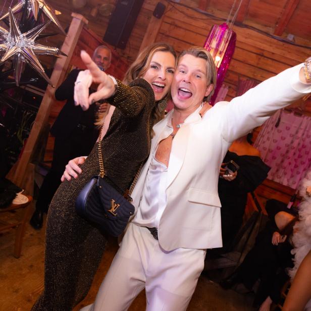 Zwei fröhliche Menschen feiern ausgelassen in eleganter Kleidung, umgeben von festlicher Dekoration und weiteren Gästen.