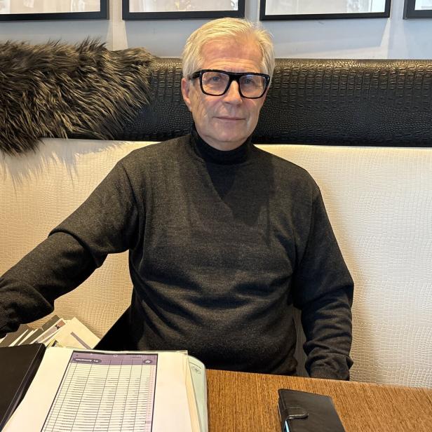 Ein älterer Mann mit Brille sitzt an einem Tisch mit Unterlagen und schaut freundlich in die Kamera.