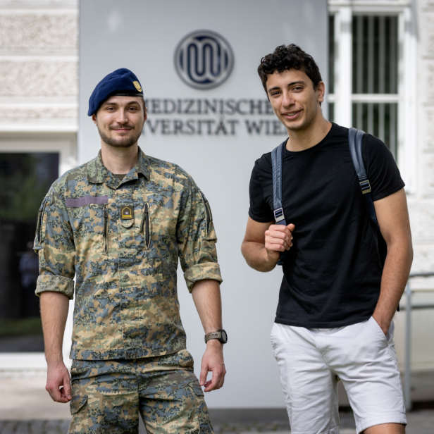 Ein Mann steht in weißer Hose, schwarzem t-Shirt und Rucksack neben einem Soldaten in Uniform, vor einem Logo der Medizinischen Universität Wien 