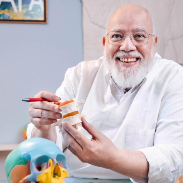 Ein Mann mit weißem Bart und Brille hält ein Knochenmodell in der Hand.