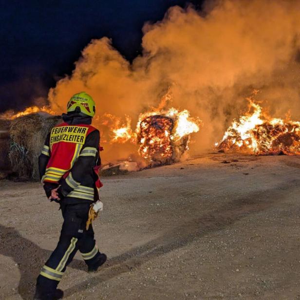 Mehrere Feuerwehrleute bekämpfen nachts einen großen Brand von brennenden Heuballen im Freien.