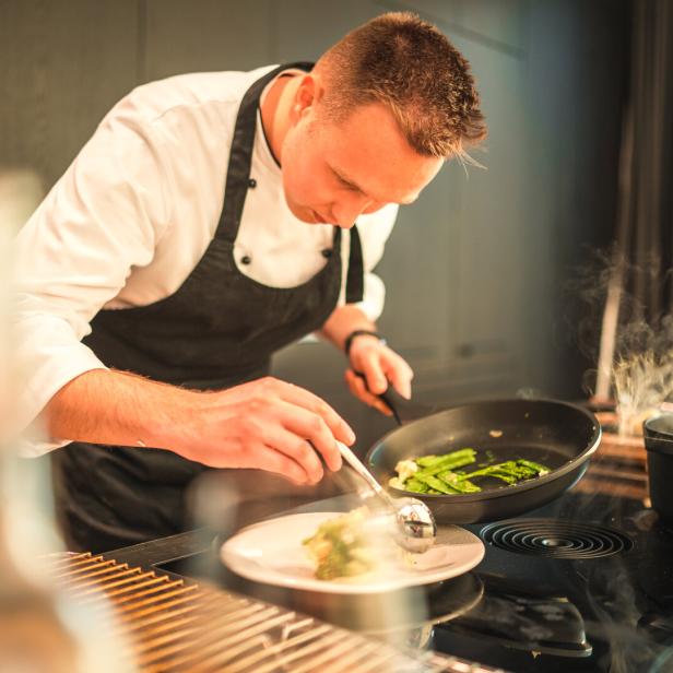 Ein Koch richtet konzentriert mit einer Kelle Gemüse aus einer Pfanne auf einem Teller an.