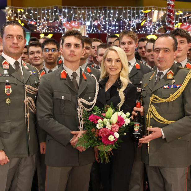 Eine Gruppe von Soldaten in Uniform steht mit einer Frau, die einen Blumenstrauß hält, vor festlicher Beleuchtung zusammen.