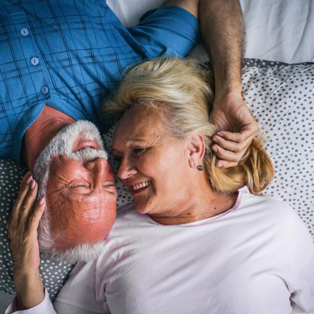 Ein älteres Paar liegt lächelnd im Bett, ihre Gesichter einander zugewandt.