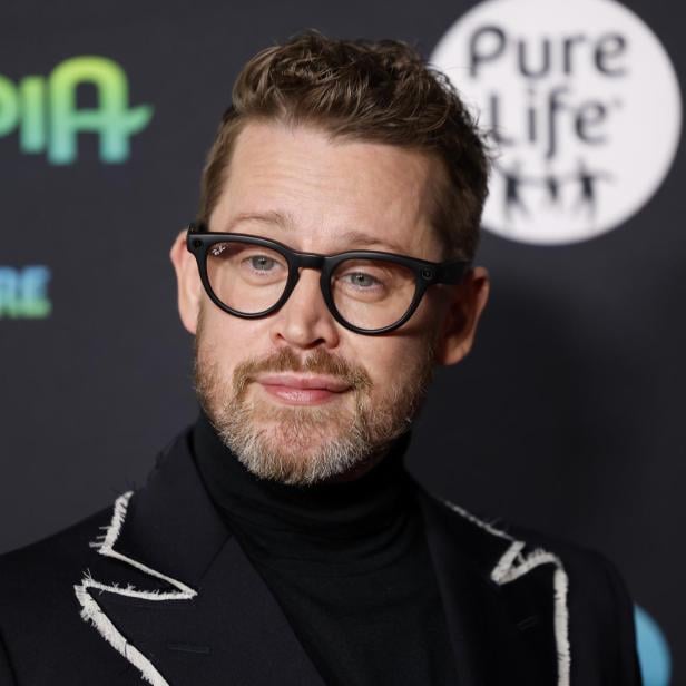 Ein Mann mit Brille, Bart und dunklem Anzug mit auffälligen Nähten steht vor einer Wand mit buntem Schriftzug.