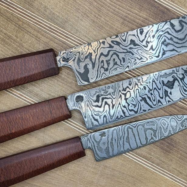 Drei handgeschmiedete Damastmesser mit markanter Musterung und Holzgriffen auf Holzuntergrund.