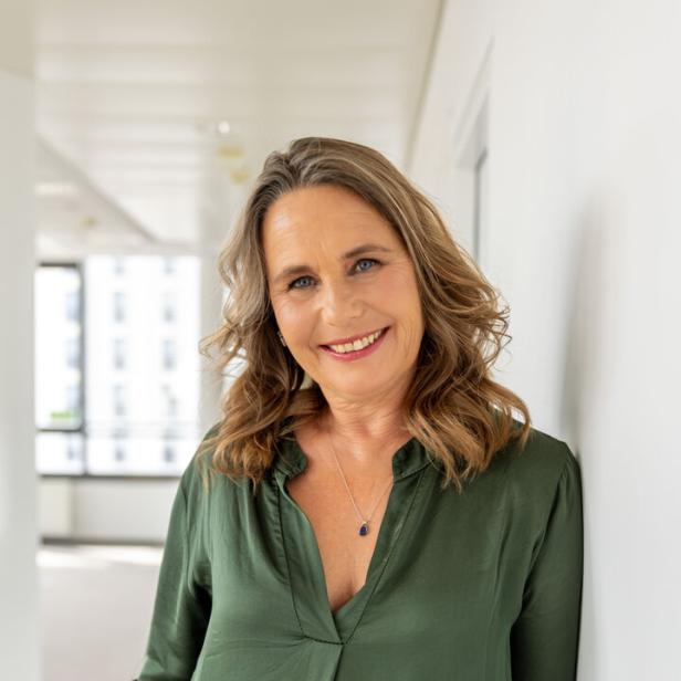 Frau mit schulterlangen, welligen Haaren und grünem Oberteil lächelt in einem hellen, modernen Raum.