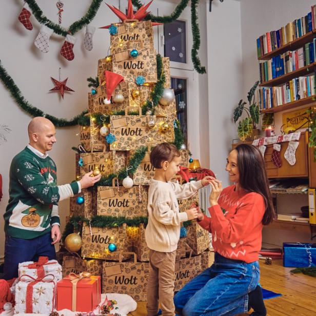 Eine Familie mit einem Kind sitzt um einen Christbaum, der aus Papiersackerl gebastelt ist, und überreicht sich Geschenke. 