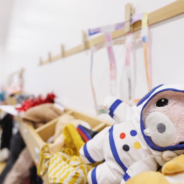 Ein rosa Kuscheltier im Astronautenanzug liegt auf einer Garderobe mit bunten Bändern und Kleidung.