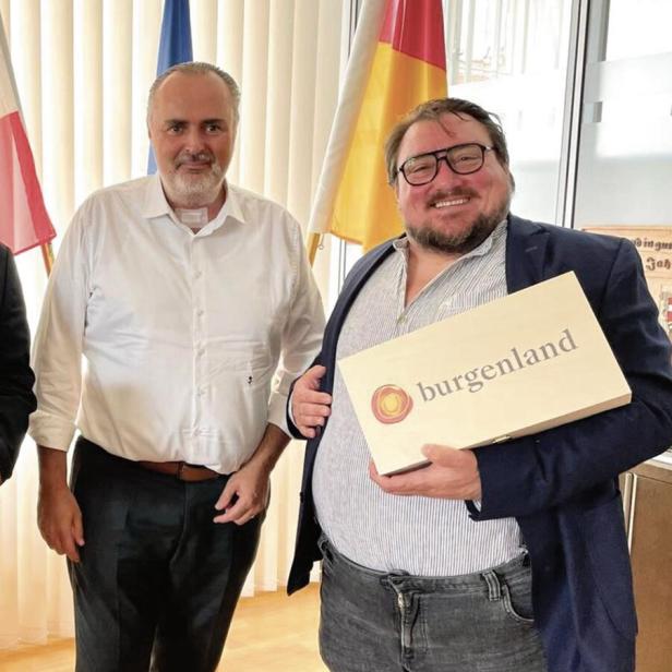 Zwei Männer stehen nebeneinander, einer hält eine Geschenkbox mit der Aufschrift „Burgenland“, im Hintergrund sind Flaggen zu sehen.