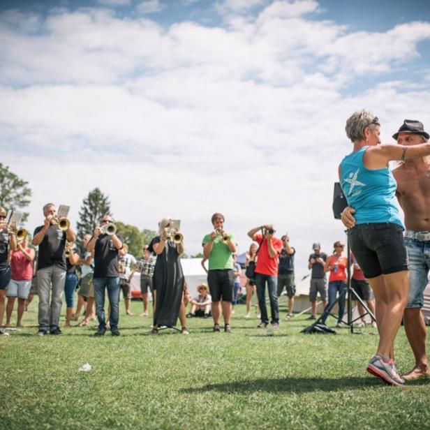 Ein Paar tanzt barfuß auf einer Wiese, während eine Blaskapelle im Hintergrund spielt und Menschen zuschauen.