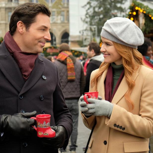 Ein Mann und eine Frau unterhalten sich lächelnd auf einem Weihnachtsmarkt und halten rote Tassen in den Händen.