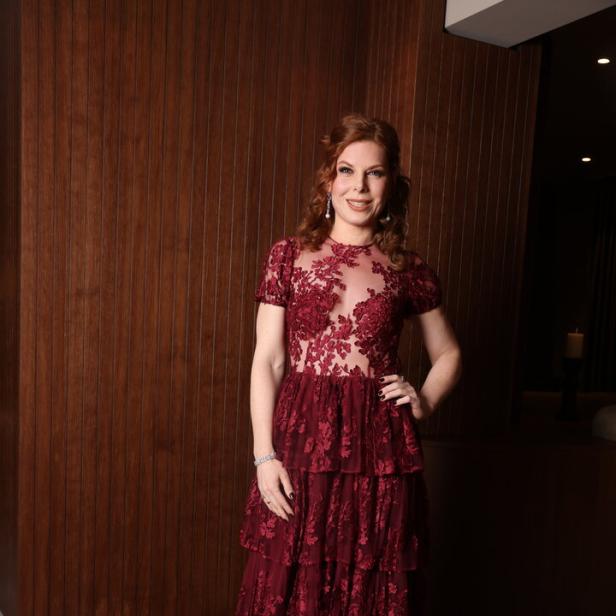 Frau mit langen, gelockten Haaren trägt ein bodenlanges, bordeauxrotes Spitzenkleid mit floralen Mustern.