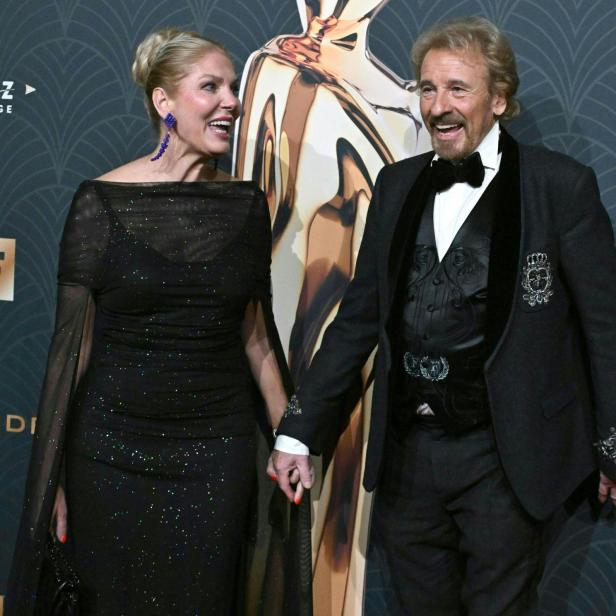 Ein elegant gekleidetes Paar lächelt und hält Händchen auf einem festlichen Event mit dunklem Hintergrund.