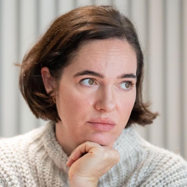 Verena Altenberger mit braunen Haaren und einem hellen Pullover stützt ihr Kinn nachdenklich ab.