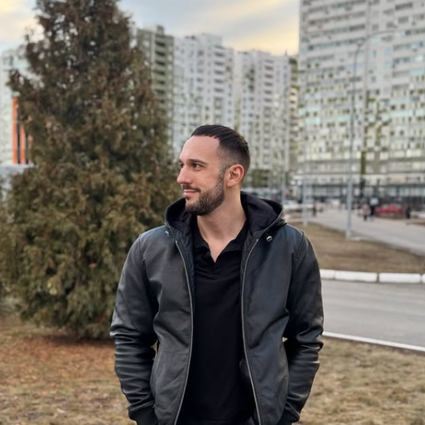 Mann steht in schwarzer Jacke und schwarzen Hosen, darüber einer Lederjacke, mit den Händen in den Hosentaschen vor einem Tannenbaum. 