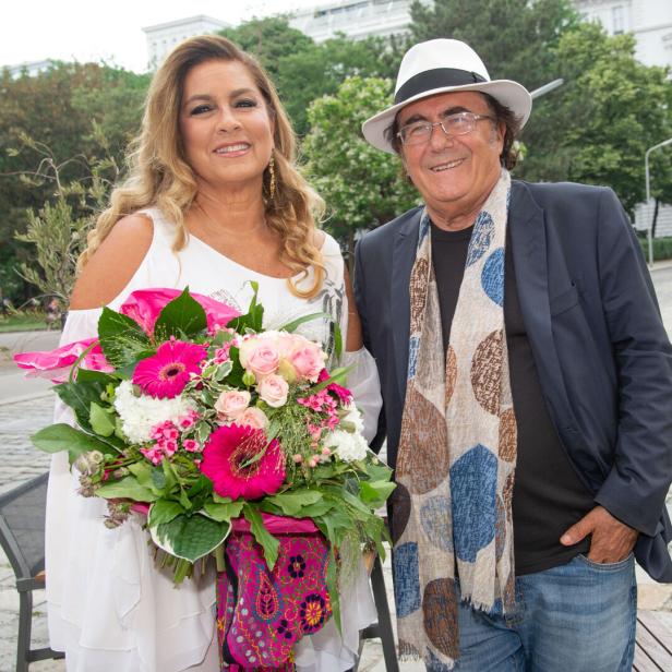 Eine Frau im weißen Kleid hält einen bunten Blumenstrauß, neben ihr steht ein Mann mit Hut und Schal, beide lächeln.