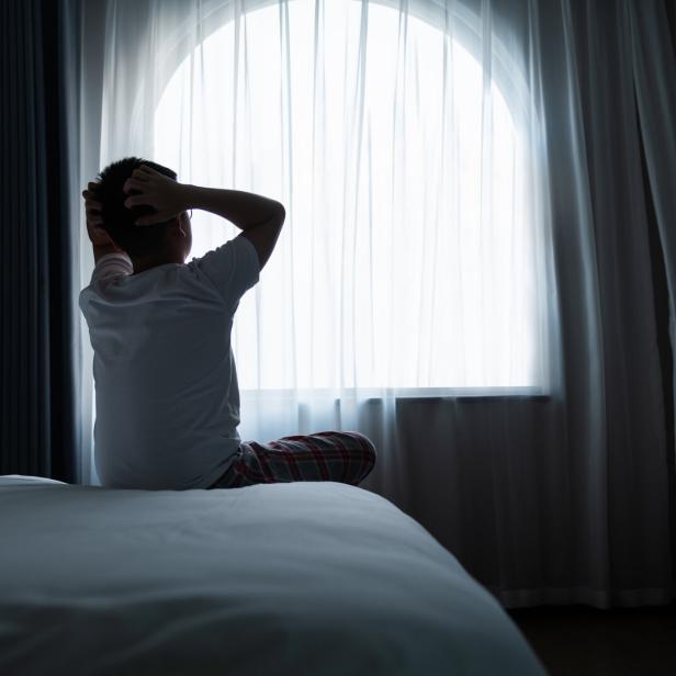 Ein Kind sitzt im Schlafanzug auf einem Bett und hält sich den Kopf vor einem hellen Fenster.