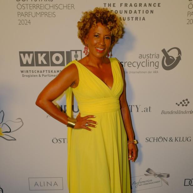 Eine Frau mit lockigen Haaren trägt ein langes, gelbes Kleid und steht lächelnd vor einer Sponsorenwand.