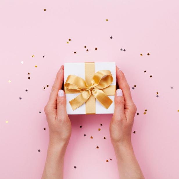 Zwei Hände halten eine weiße Geschenkbox mit goldener Schleife auf rosa Hintergrund mit kleinen Sternkonfetti.