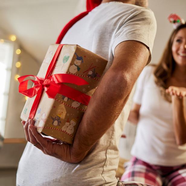 Ein Mann versteckt ein Geschenk mit roter Schleife hinter dem Rücken, während eine Frau erwartungsvoll lächelt.