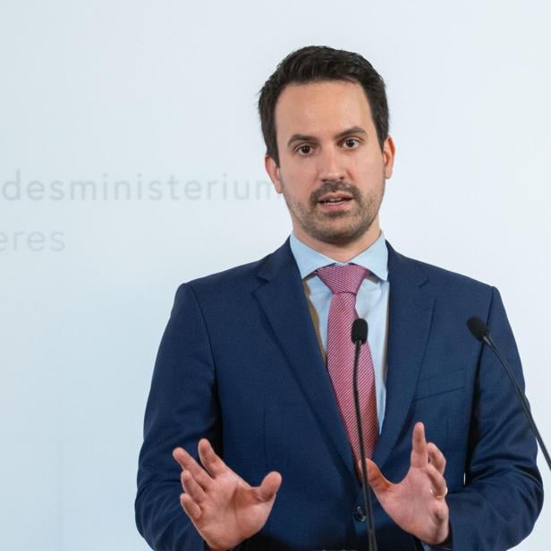 Ein Mann im Anzug spricht an zwei Mikrofonen und gestikuliert mit den Händen vor hellem Hintergrund.