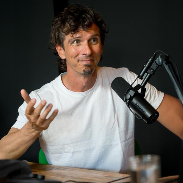 Mann mit weißem T-Shirt gestikuliert mit einer Hand vor einem Mikrofon an einem Tisch mit Laptop und Glas.