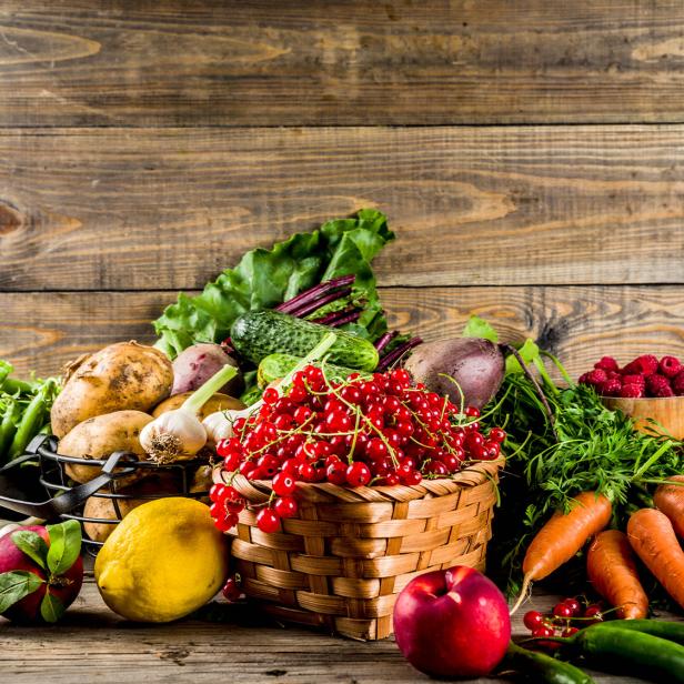 Korb mit roten Johannisbeeren, umgeben von Karotten, Äpfeln, Kartoffeln, Gurken, Bananen, Erbsen und anderem Gemüse auf Holztisch.