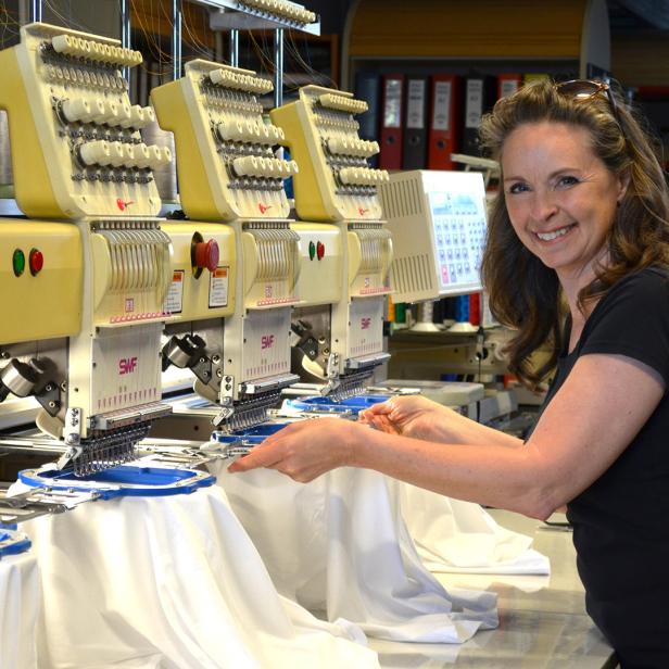 Frau mit dunklem T-Shirt bedient mehrere industrielle Stickmaschinen mit weißen Stoffen in einer Werkstatt.