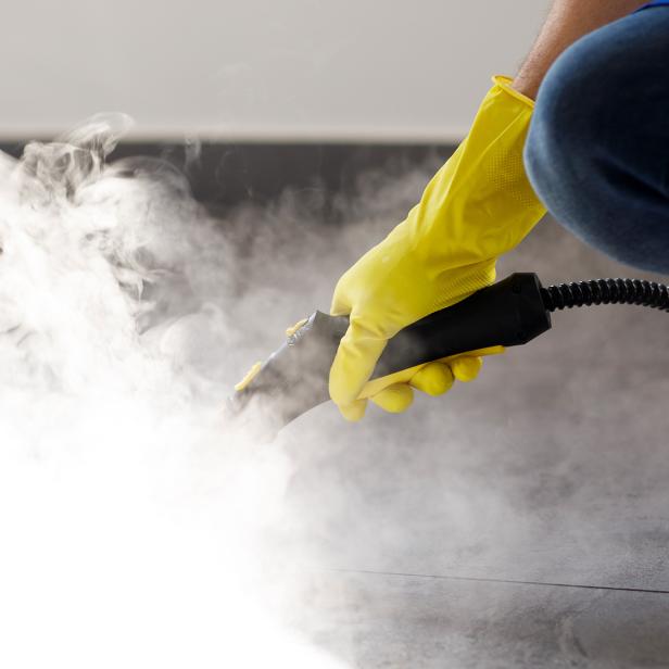 Hand mit gelbem Handschuh reinigt Bodenfliesen mit einem Dampfreiniger.