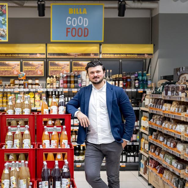 Ein Mann steht in einem Supermarkt neben Getränkekisten, umgeben von regionalen Produkten und Regalen mit Lebensmitteln.