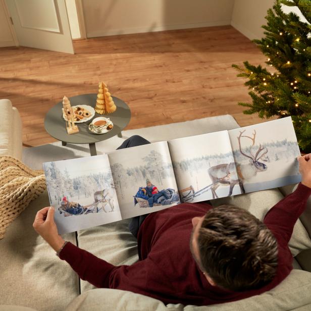 Mann betrachtet auf dem Sofa ein Fotobuch mit Wintermotiven neben einem geschmückten Weihnachtsbaum.