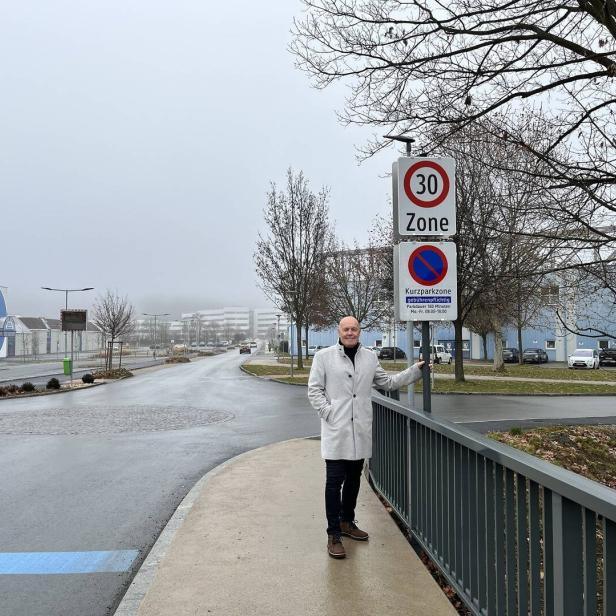 Ein Mann im hellen Mantel steht an einem Geländer neben Verkehrsschildern auf einer nassen, leeren Straße mit kahlen Bäumen.