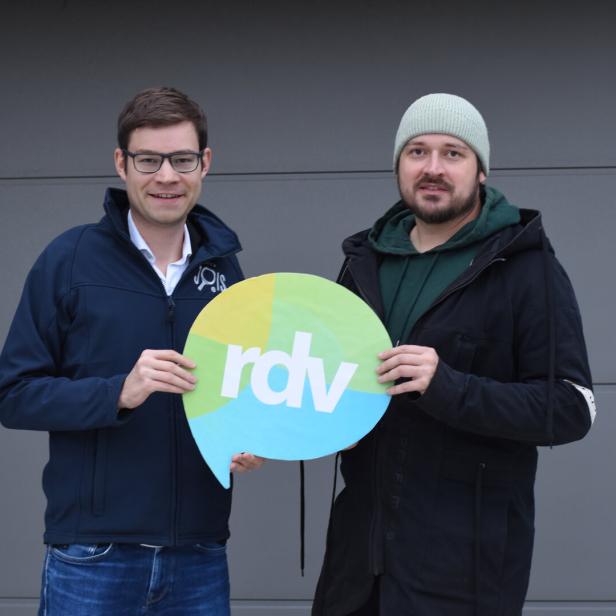 Zwei Männer stehen nebeneinander und halten gemeinsam ein rundes Schild mit der Aufschrift „rdv“.