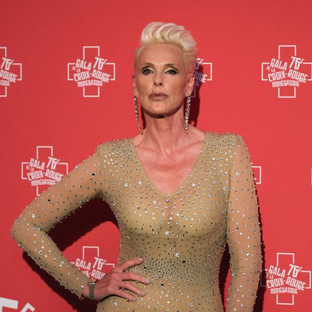 Brigitte Nielson mit blonden, kurzen Haaren trägt ein glitzerndes Kleid vor rotem Hintergrund.