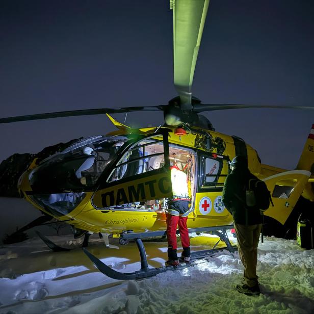 Ein gelber Rettungshubschrauber steht nachts auf verschneitem Boden, zwei Personen stehen beleuchtet daneben.