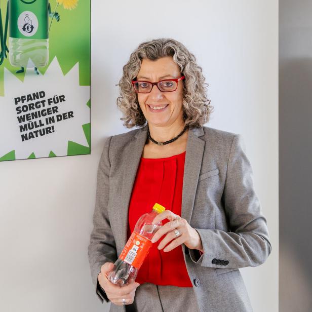 Eine Frau mit grauen Locken und rotem Oberteil hält eine Pfandflasche vor einem Plakat zum Thema Recycling.
