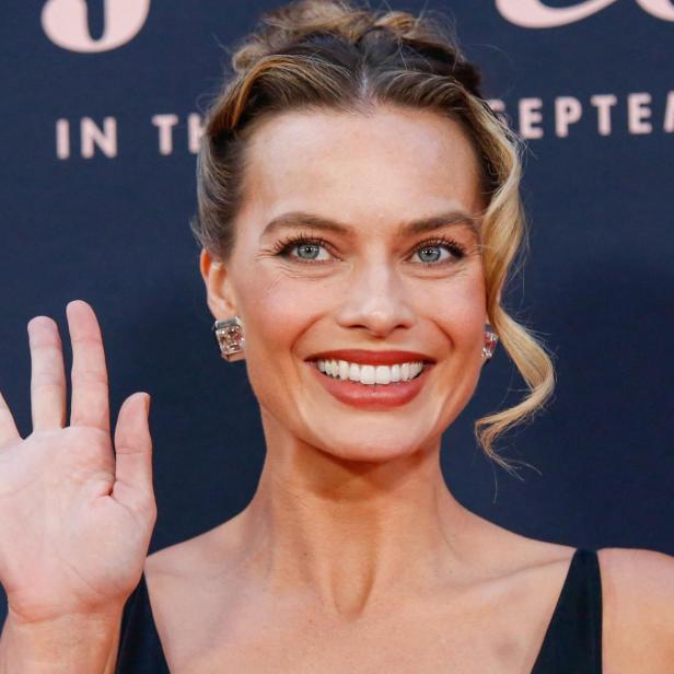 Eine Frau mit hochgesteckten Haaren lächelt und winkt mit der rechten Hand vor dunklem Hintergrund.