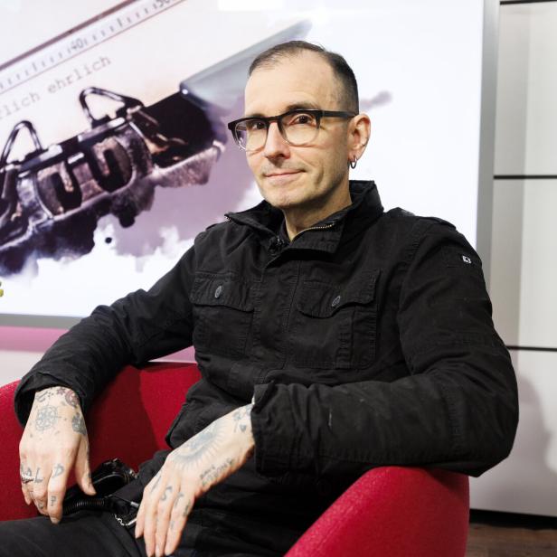 Ein Mann mit Brille und Tattoos sitzt in einem roten Sessel, im Hintergrund eine Schreibmaschine und eine pinke Orchidee.