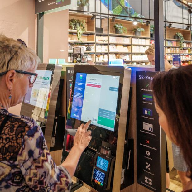 Zwei Personen bedienen eine Selbstbedienungskasse in einem modernen Geschäft mit Regalen im Hintergrund.